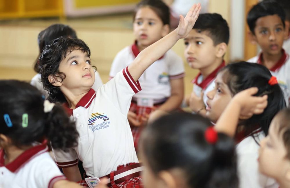 yoga Large A student in a classroom looking upwards with her hand raised, as if showing something to her classmates while they watch attentively.